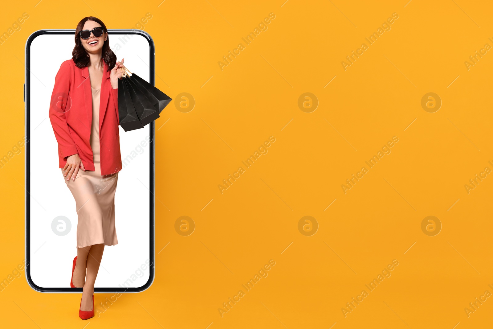 Image of Online shopping. Happy woman with paper bags walking out from smartphone on orange background, space for text