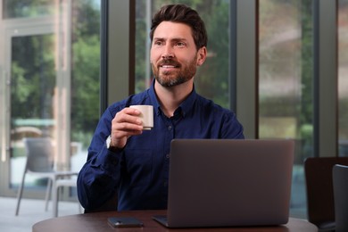 Photo of Man with cup of drink and laptop at table in cafe