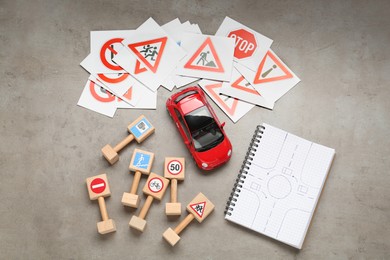 Photo of Many different road signs, notebook with sketch of roundabout and toy car on grey table, flat lay. Driving school