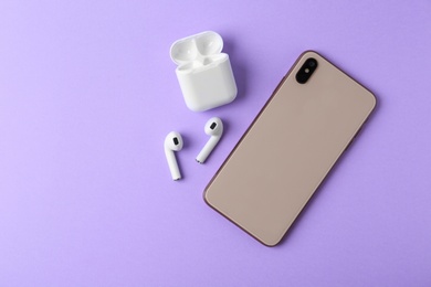 Photo of Wireless earphones, mobile phone and charging case on violet background, flat lay. Space for text