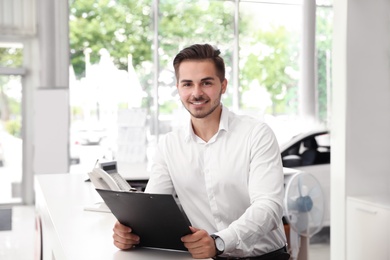 Photo of Young salesman with clipboard at desk in car salon