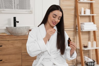 Happy young woman with bottle applying essential oil onto hair in bathroom