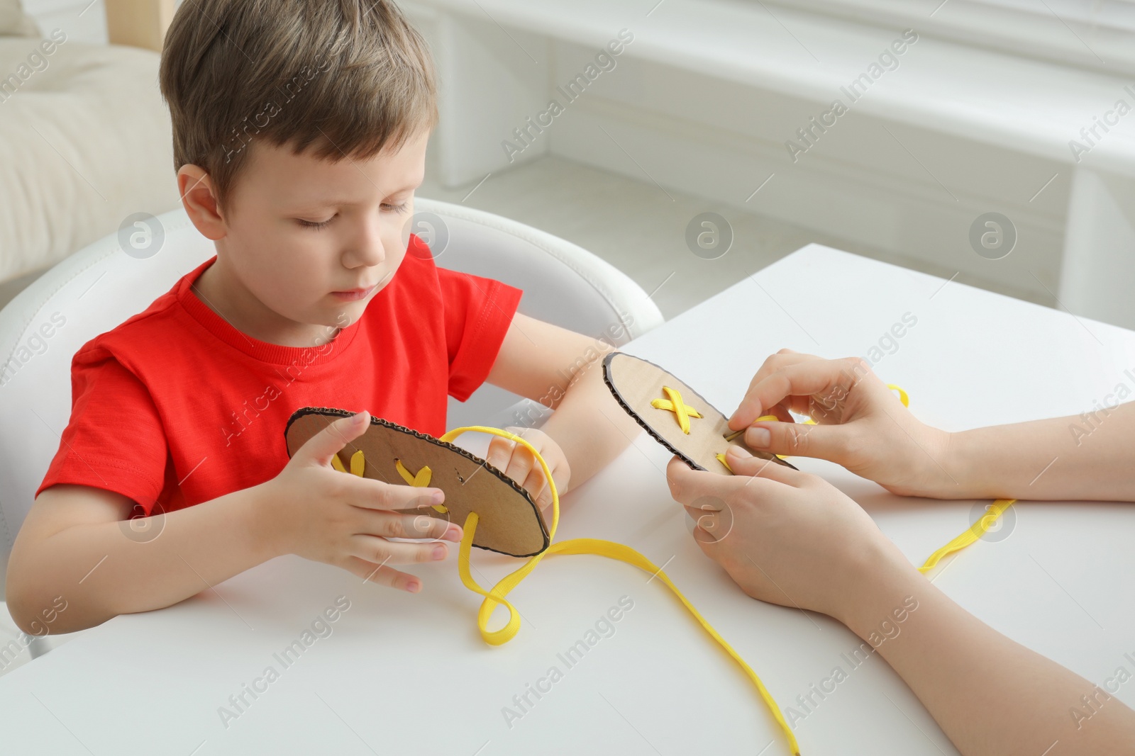 Photo of Mother teaching son to tie shoe laces using training cardboard template at white table, closeup