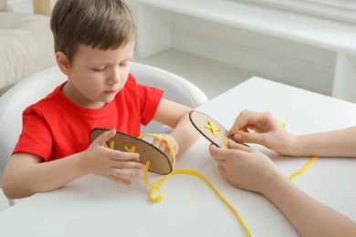 Mother teaching son to tie shoe laces using training cardboard template at white table, closeup