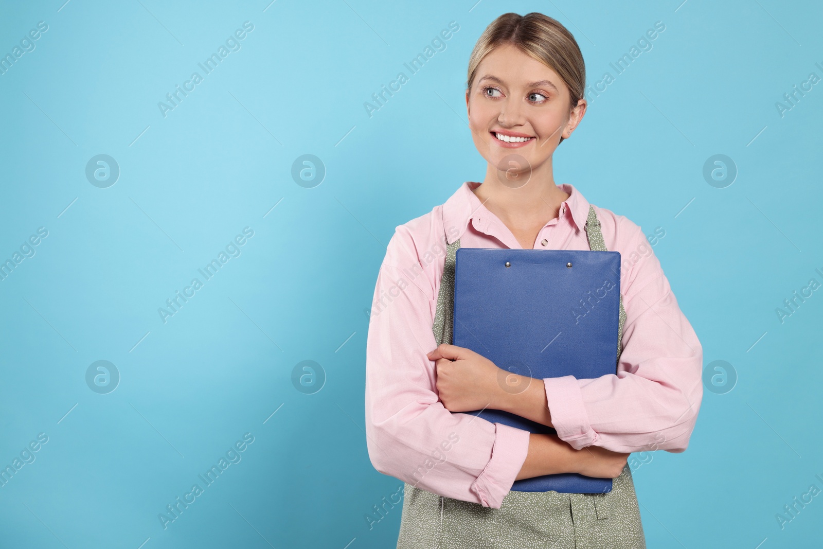 Photo of Beautiful young woman in clean apron with clipboard on light blue background. Space for text