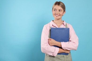 Photo of Beautiful young woman in clean apron with clipboard on light blue background. Space for text