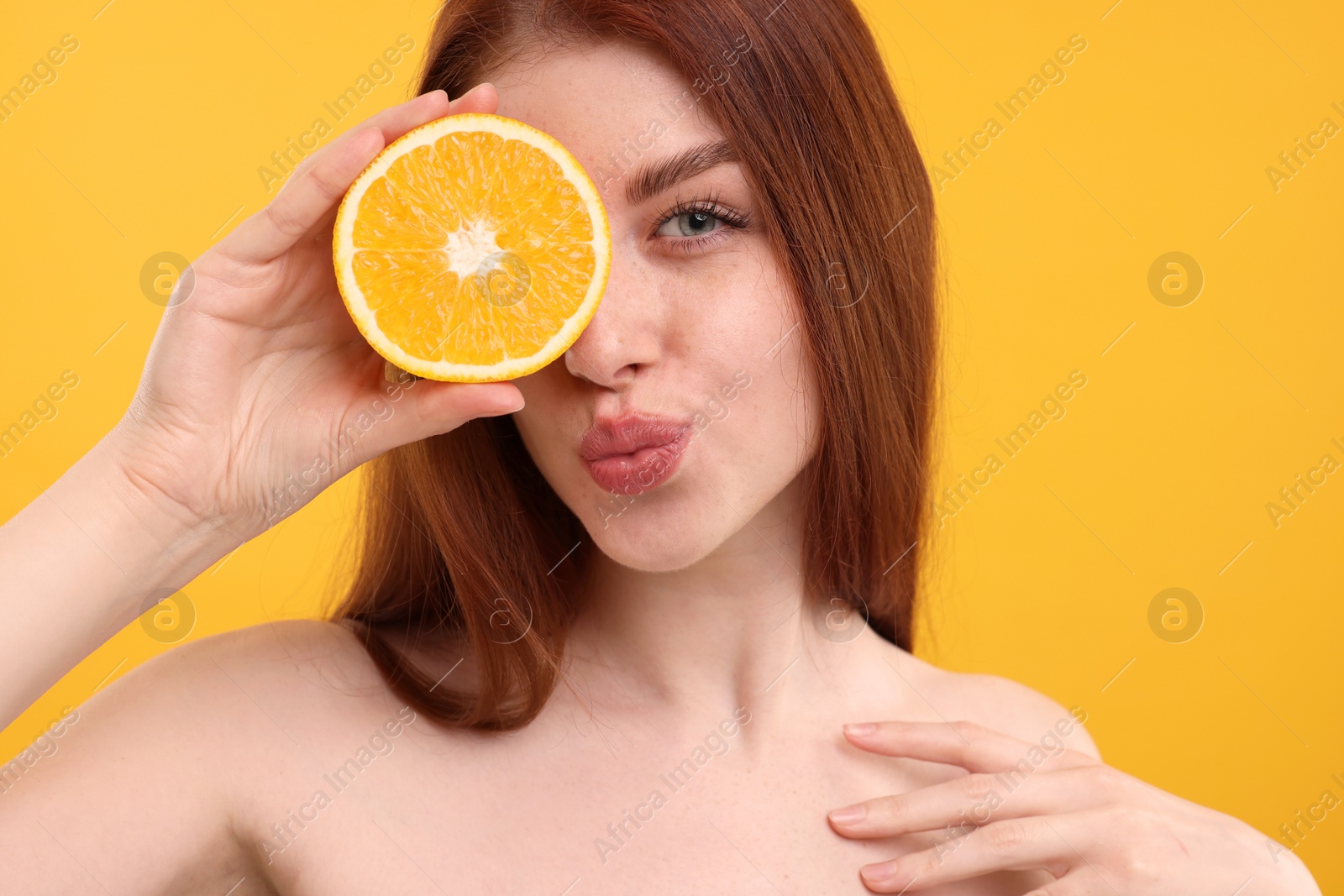 Photo of Beautiful woman with freckles covering eye with half of orange and sending air kiss on yellow background