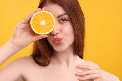 Beautiful woman with freckles covering eye with half of orange and sending air kiss on yellow background