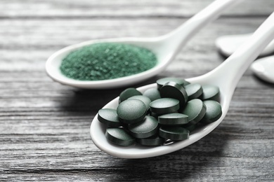 Photo of Spoons of spirulina pills and powder on wooden table, closeup