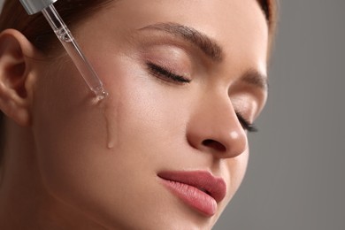 Photo of Beautiful young woman applying cosmetic serum onto her face on grey background, closeup