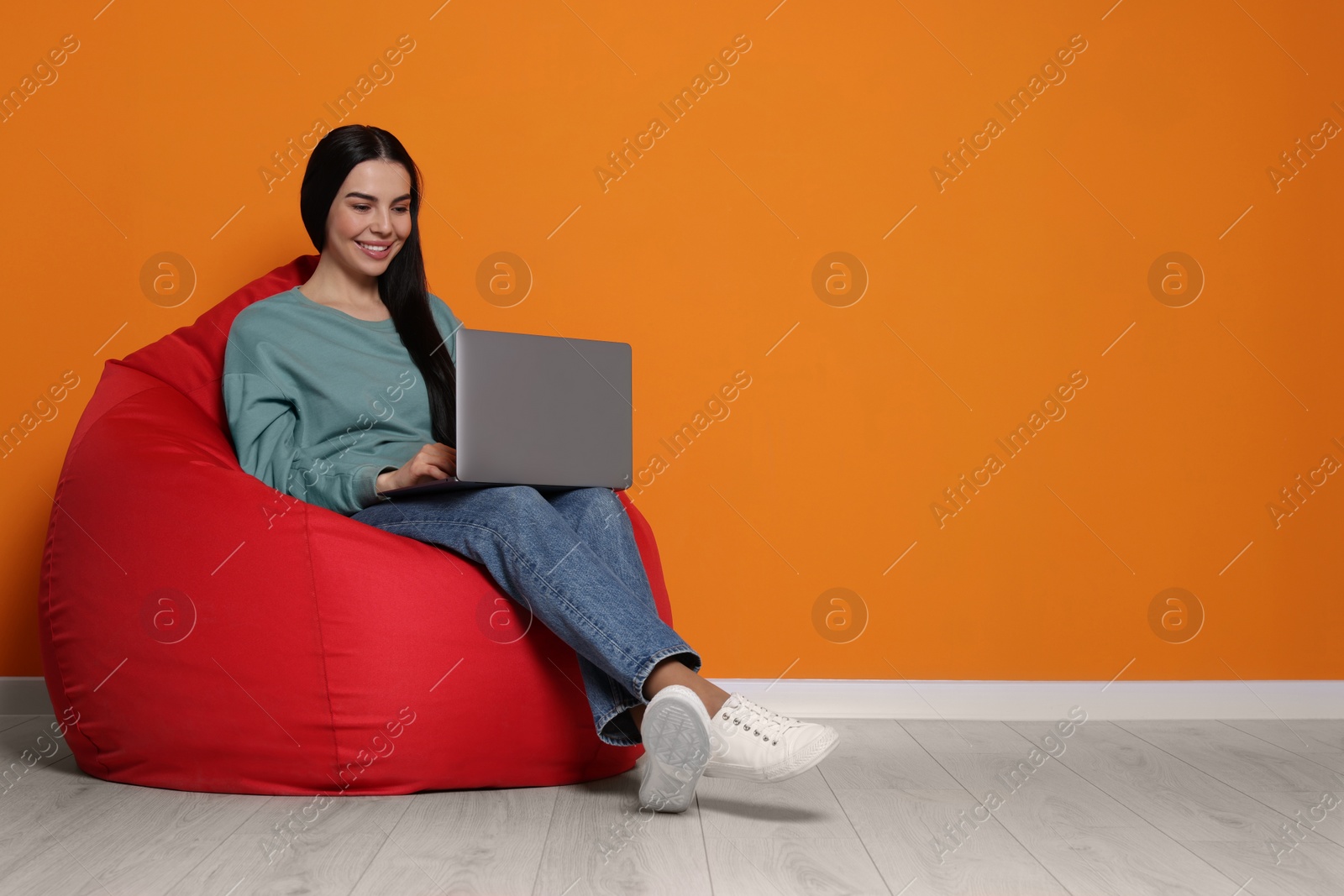 Photo of Happy woman with laptop sitting on beanbag chair near orange wall, space for text