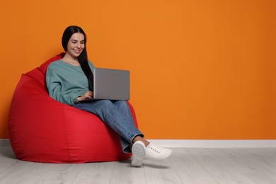 Happy woman with laptop sitting on beanbag chair near orange wall, space for text