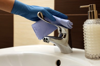 Photo of Woman in gloves cleaning faucet of bathroom sink with rag, closeup