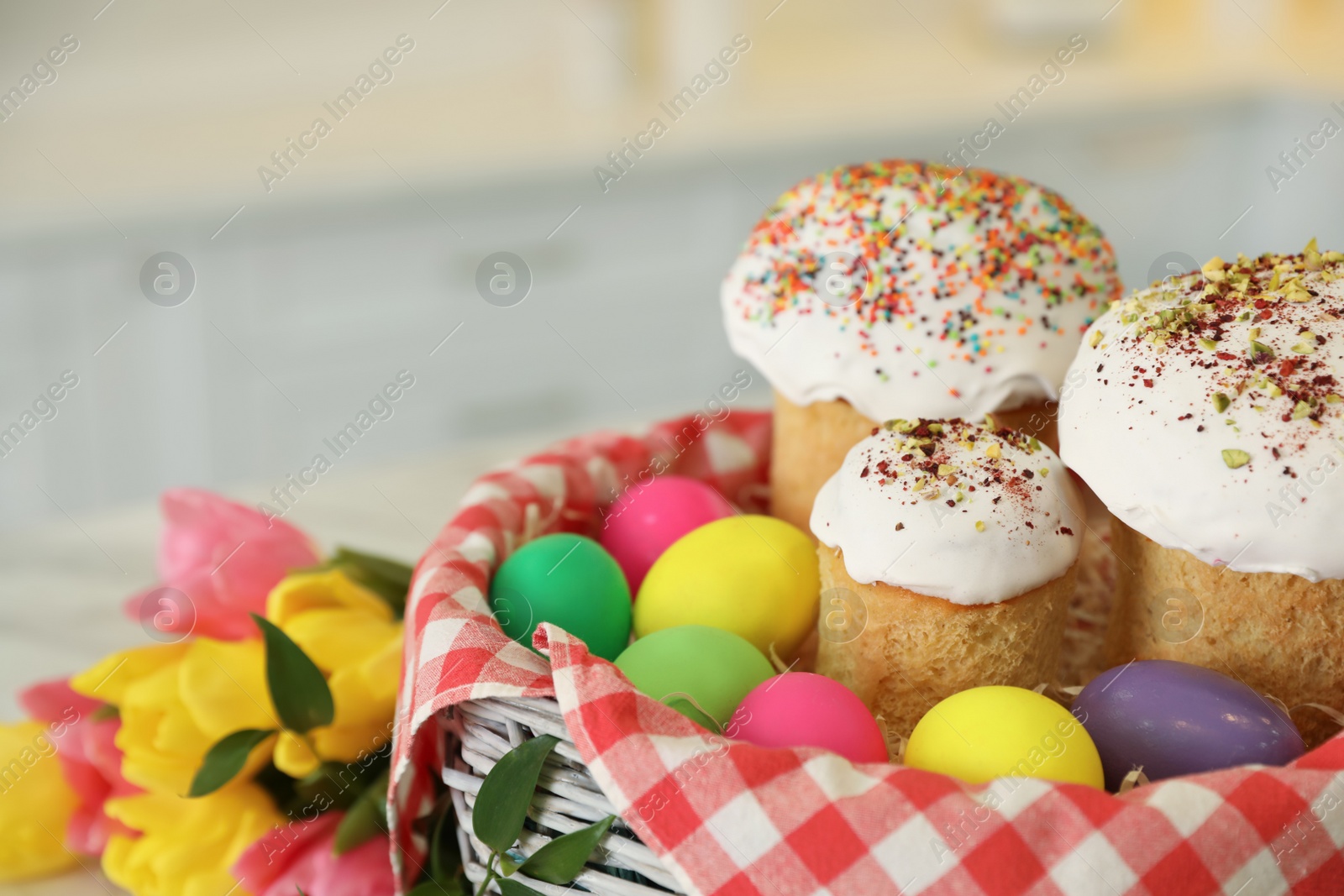 Photo of Basket with traditional Easter cakes, dyed eggs and flowers on blurred background, closeup