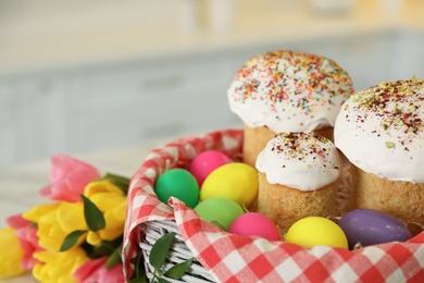 Basket with traditional Easter cakes, dyed eggs and flowers on blurred background, closeup