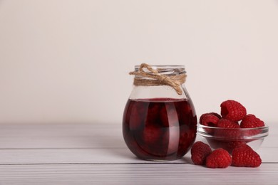 Photo of Jar of tasty canned raspberry jam and fresh berries in glass bowl on white wooden table, space for text