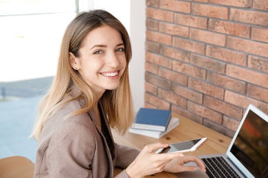 Young businesswoman with smartphone using laptop at table in office