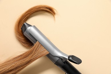 Curling iron with hair lock on beige background, closeup. Space for text