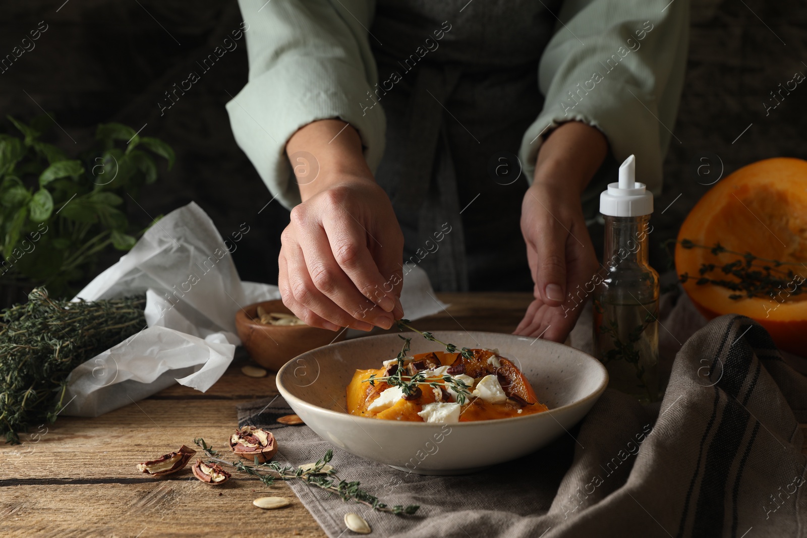 Photo of Woman adding aromatic thyme onto freshly baked pumpkin slices with cheese and pecans at wooden table, closeup