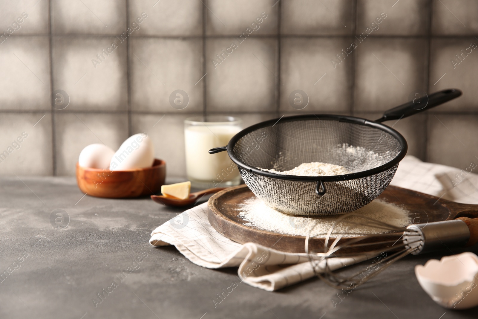 Photo of Flour in sieve and other ingredients for dough on grey textured table, closeup. Space for text