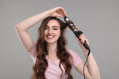 Photo of Beautiful young woman using curling hair iron on grey background