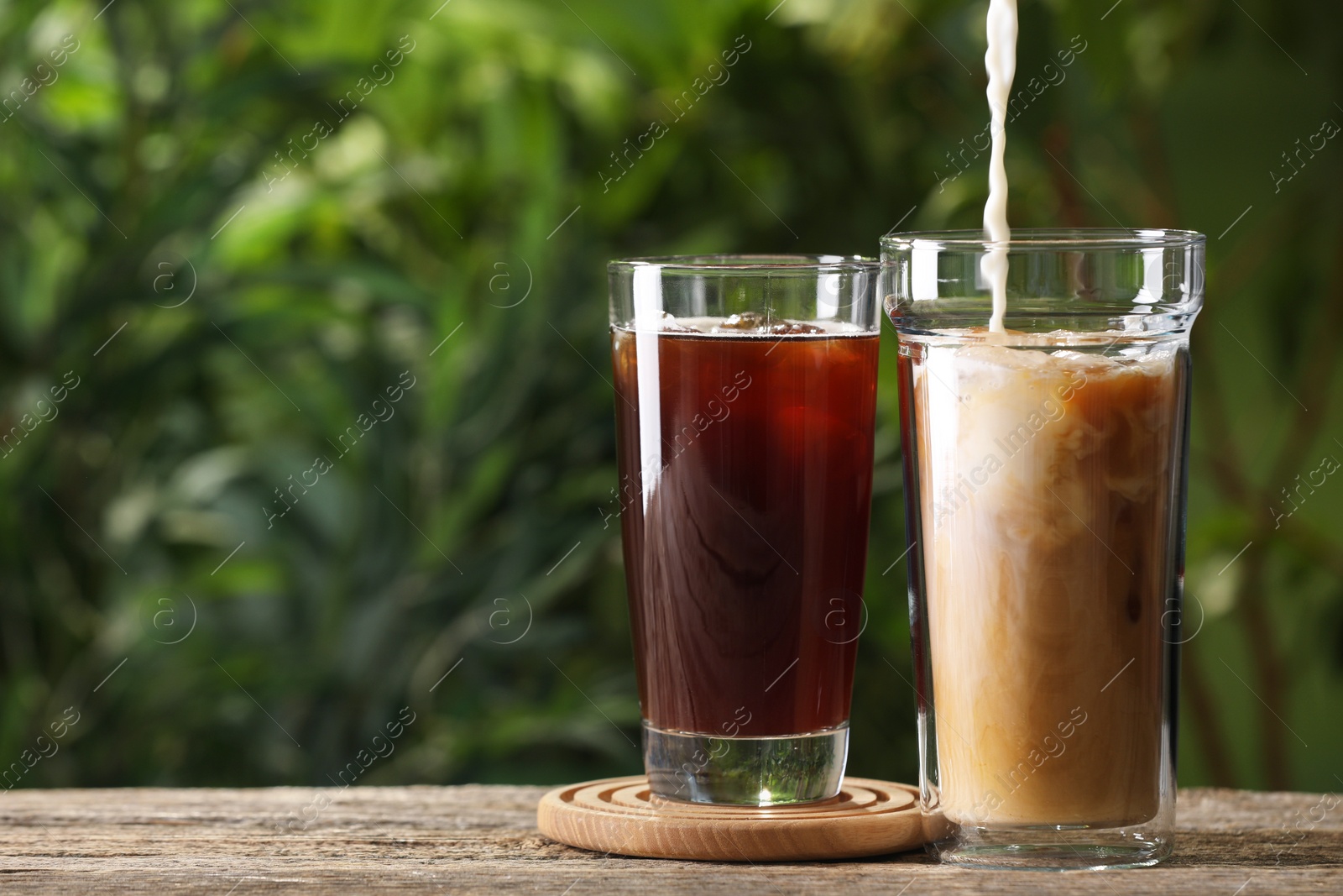 Photo of Pouring milk into iced coffee on wooden table outdoors. Space for text