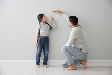 Father measuring little girl's height near light grey wall indoors