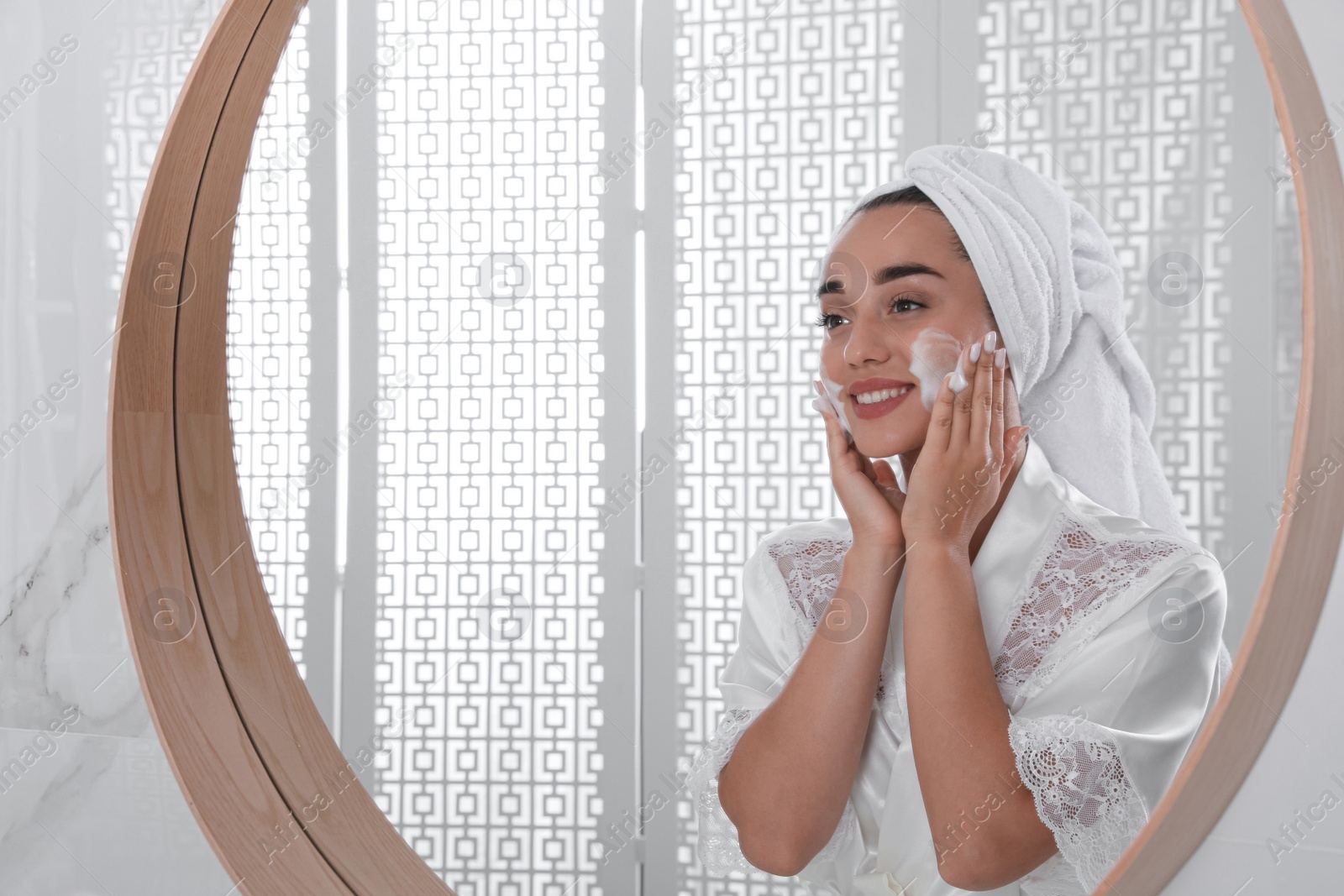 Photo of Beautiful young woman applying cleansing foam onto face near mirror in bathroom. Skin care cosmetic