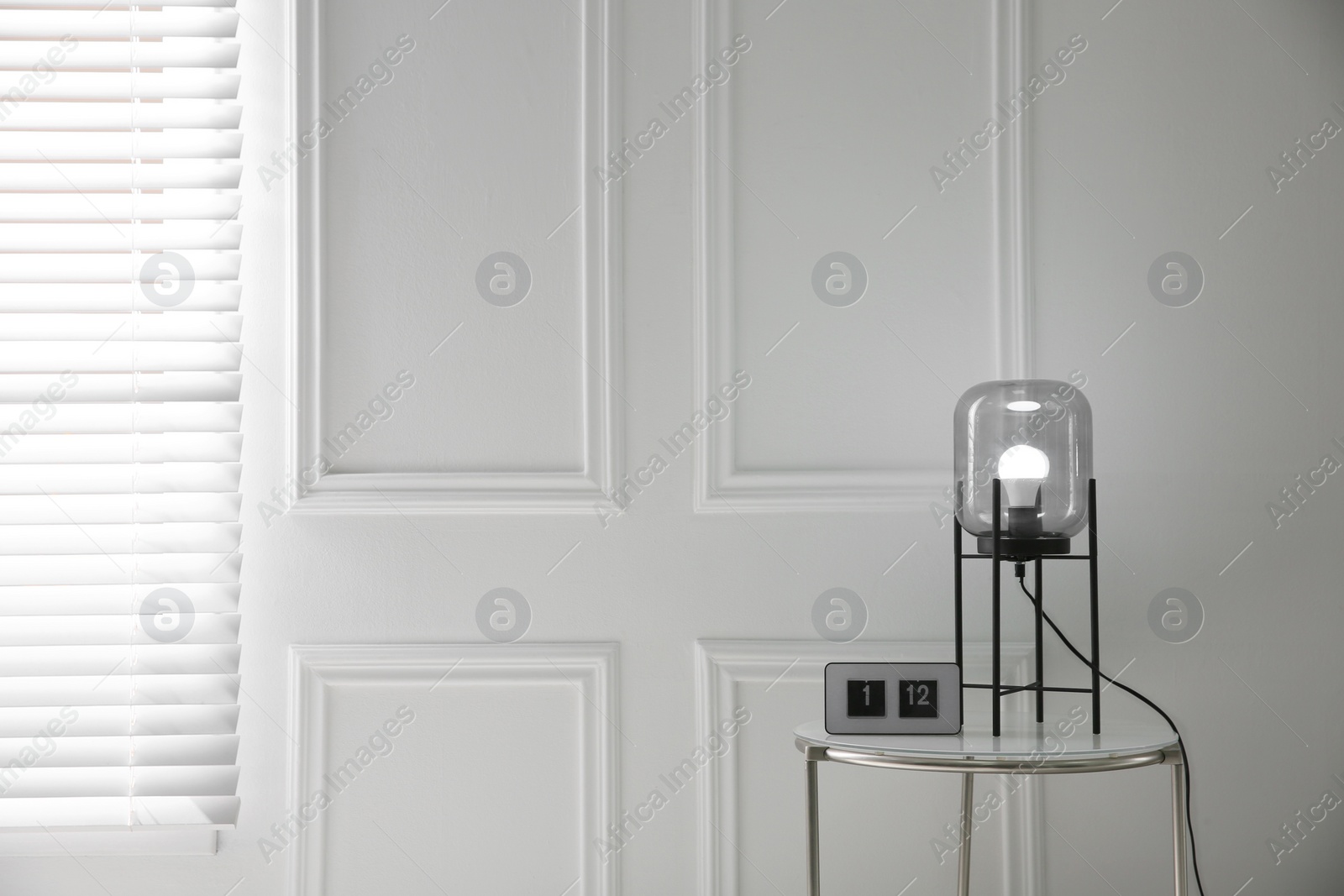 Photo of Table with clock and modern lamp near white wall in room, space for text. Interior element