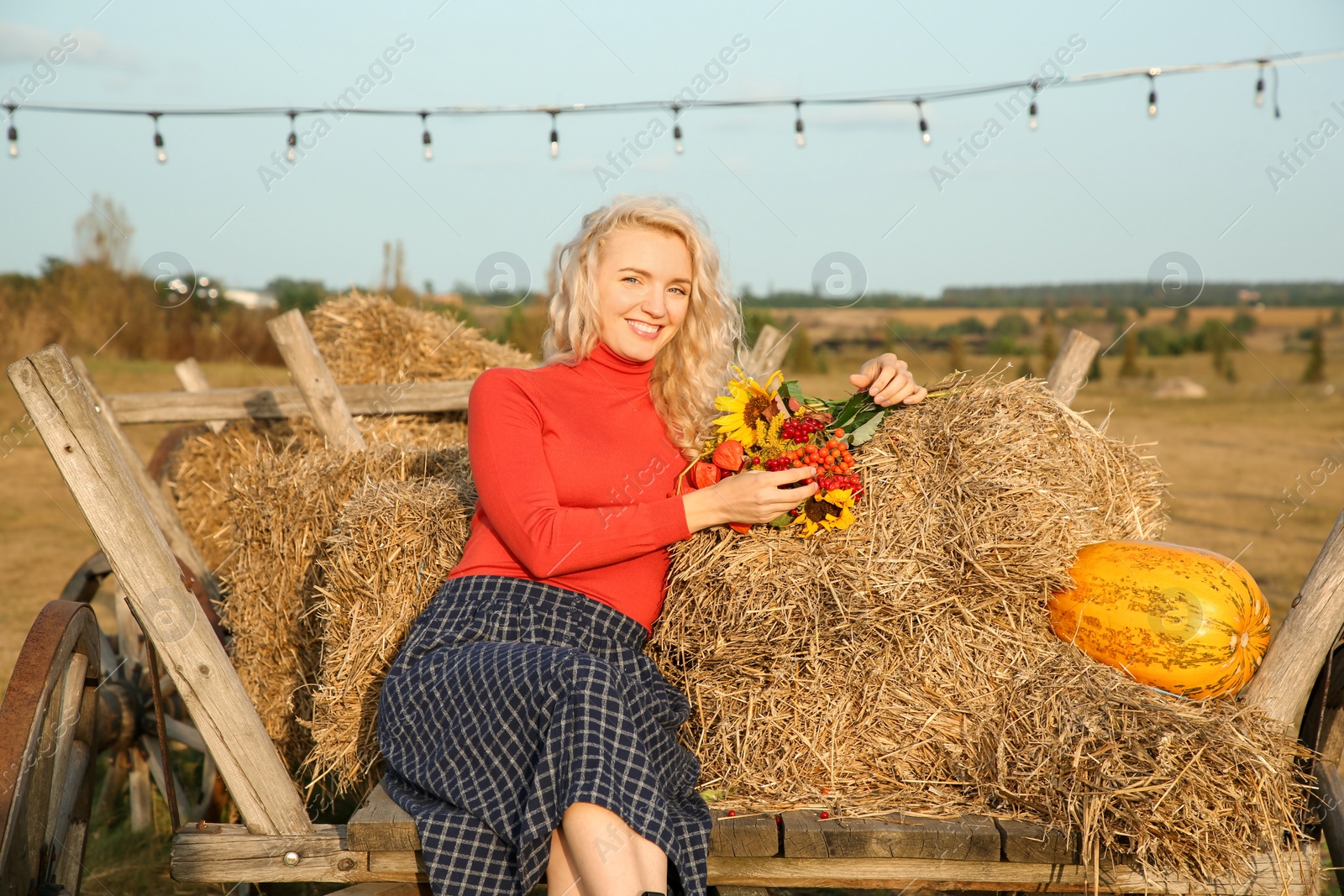 Photo of Beautiful woman with bouquet sitting on wooden cart with pumpkin and hay in field. Autumn season