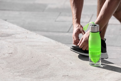 Photo of Sporty man tying shoelaces near bottle of water outdoors on sunny day. Space for text