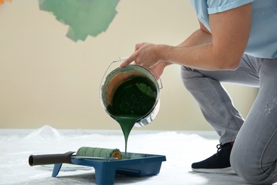 Photo of Young man pouring paint from bucket into tray indoors, closeup. Home repair