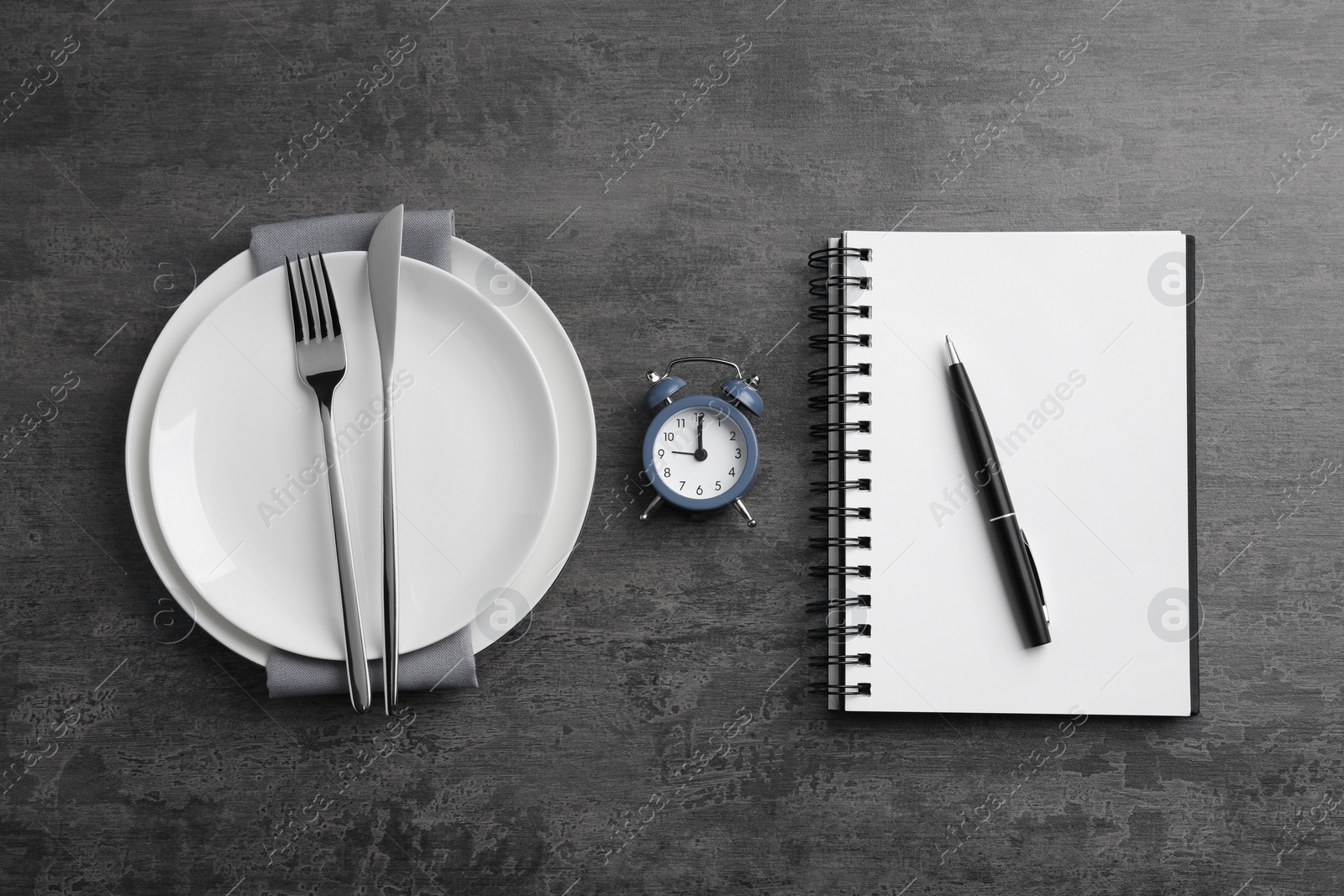 Photo of Business lunch concept. Plates, cutlery, alarm clock, notebook and pen on gray textured background, flat lay