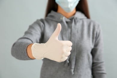 Woman in protective face mask and medical gloves showing thumb up gesture on grey background, closeup