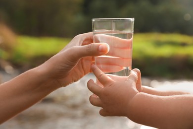 Photo of Mother giving her daughter glass of fresh water near stream on sunny day, closeup
