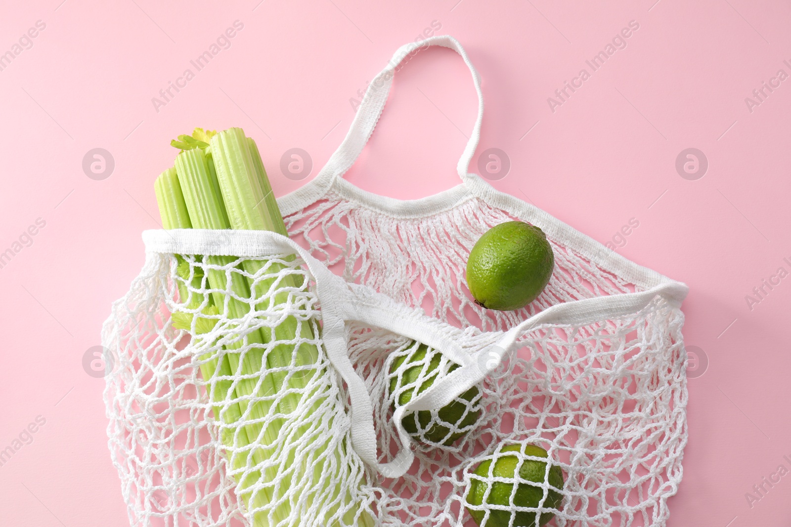 Photo of String bag with celery and limes on pink background, top view