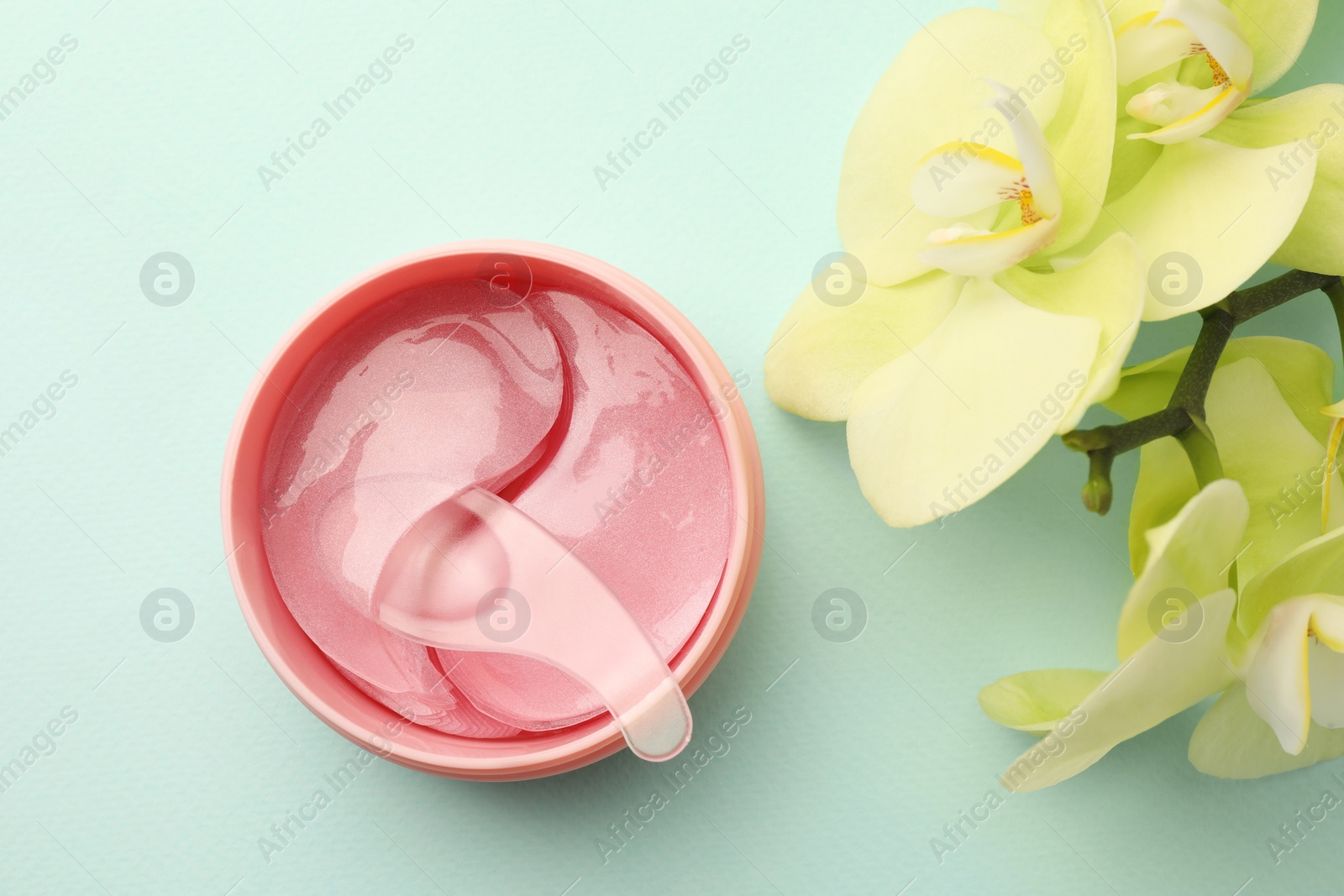 Photo of Jar of under eye patches with spoon and flowers on turquoise background, flat lay. Cosmetic product