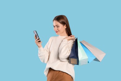 Photo of Beautiful young woman with shopping bags looking at smartphone on light blue background. Big sale
