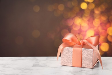 Image of Beautiful gift box on white marble table against blurred festive lights, bokeh effect. Space for text