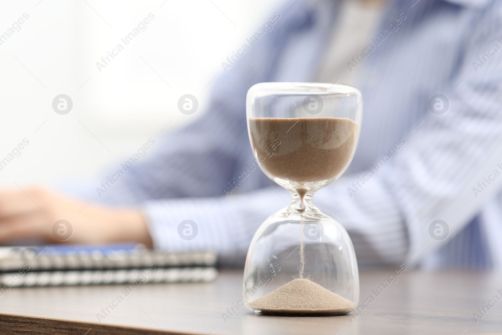 Photo of Hourglass with flowing sand on desk. Woman working indoors, selective focus