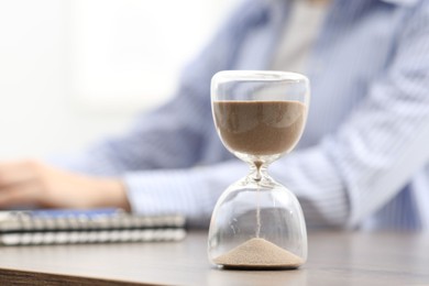 Hourglass with flowing sand on desk. Woman working indoors, selective focus