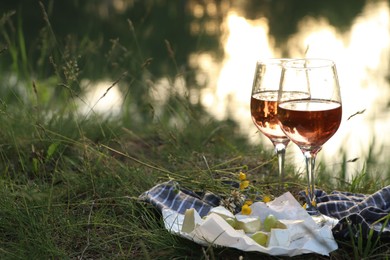 Photo of Glasses of delicious rose wine, cheese and grapes on picnic blanket near lake, space for text