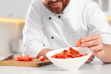 Photo of Closeup of professional chef adding tomato into bowl with delicious spaghetti at marble table in kitchen, focus on food