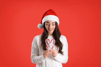 Beautiful woman in Santa Claus hat making heart with candy canes on red background