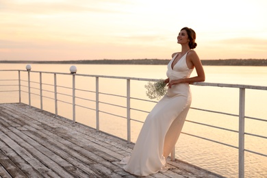 Gorgeous bride in beautiful wedding dress with bouquet near river on sunset. Space for text