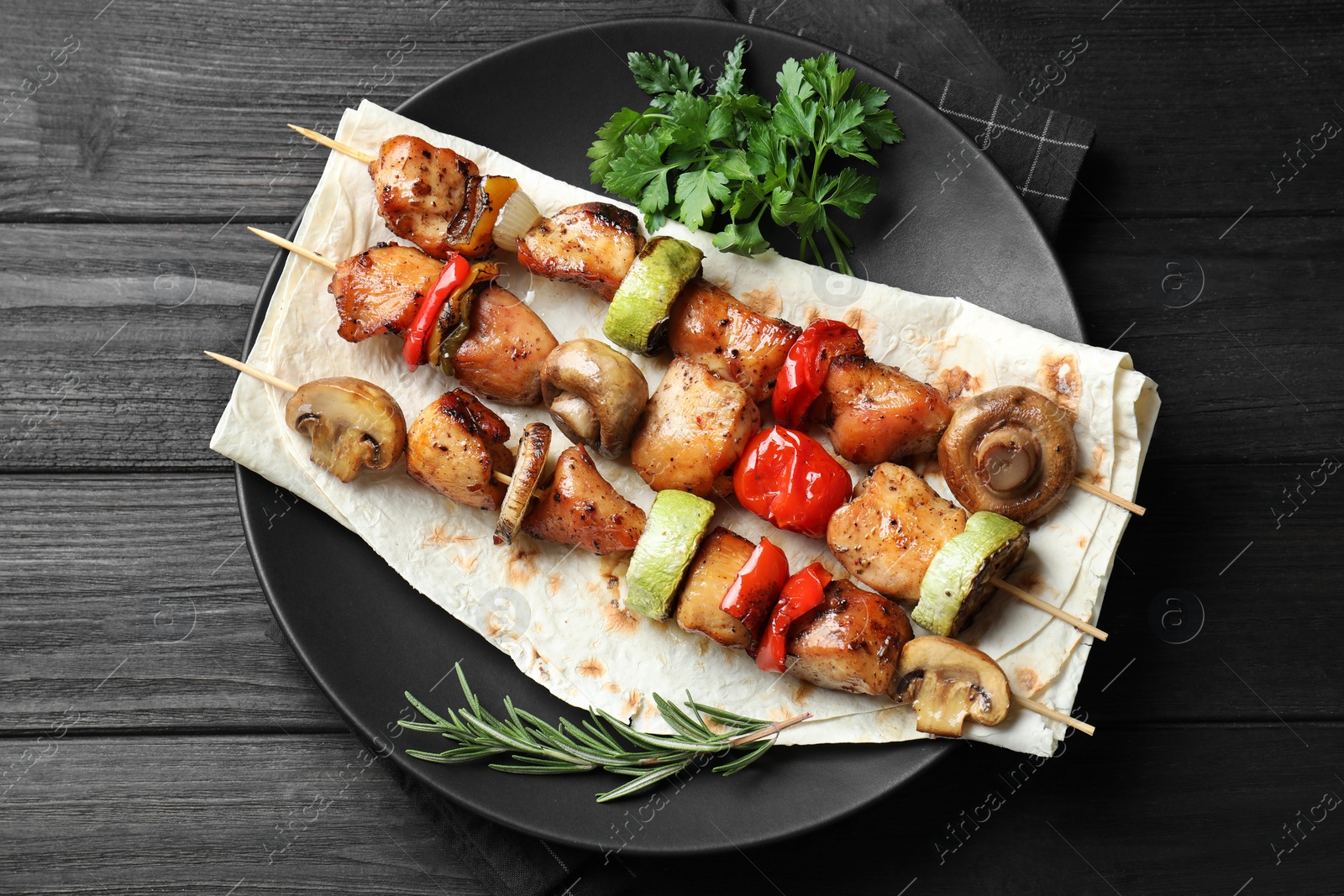 Photo of Delicious shish kebabs with vegetables and spices on black wooden table, top view
