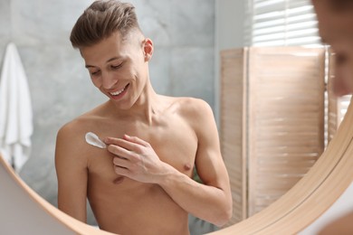 Photo of Handsome man applying moisturizing cream onto his shoulder in bathroom. Space for text