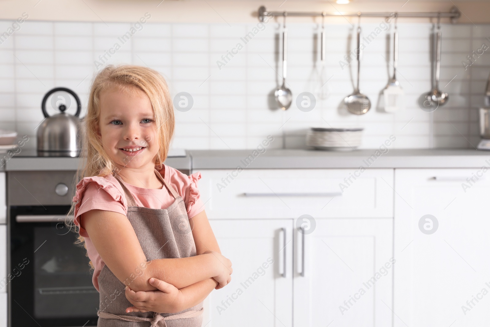 Photo of Cute little girl wearing apron near oven in kitchen. Space for text