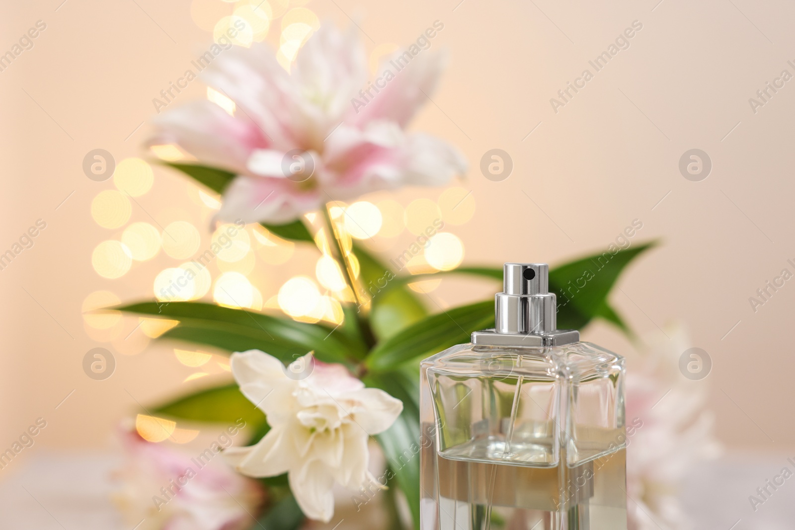 Photo of Bottle of perfume and beautiful lily flowers against beige background with blurred lights, closeup