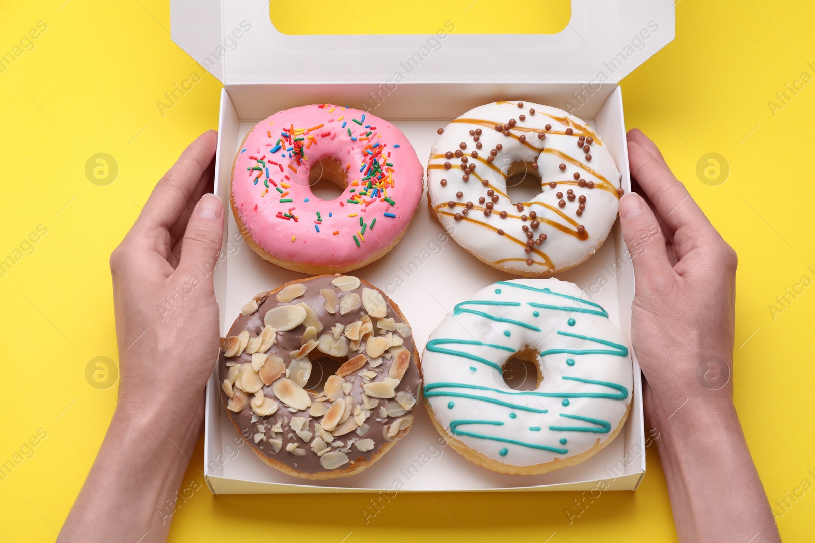 Photo of Woman holding box with tasty glazed donuts on yellow background, top view